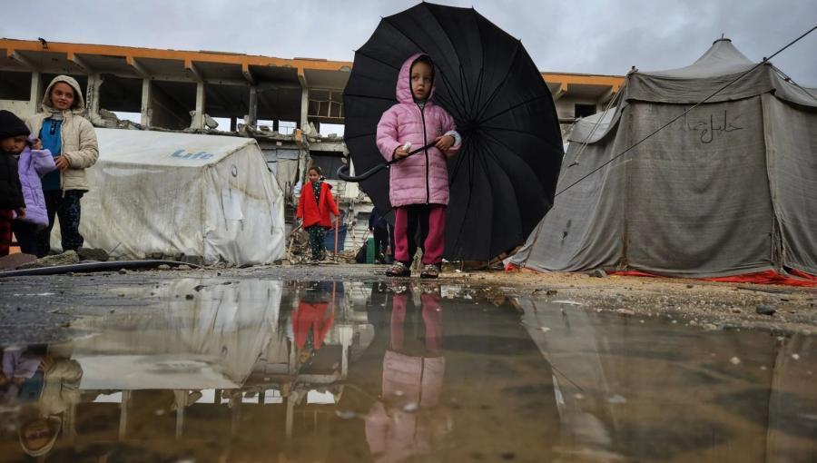 طفلة غزية تحمل مظلة أكبر من حجمها، تحاول الاحتماء بها من المطر وتنعكس صورتها في بركة ماء أمامها، بالقرب من خيام مهترئة ومبانٍ مدمرة جراء حرب الإبادة الإسرائيلية في قطاع غزة | المصدر: TRT