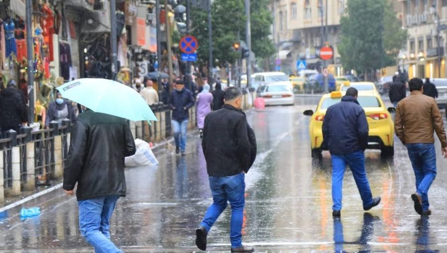 مشهد شتوي من أحد شوارع العاصمة عمان، حيث يسير الناس تحت المطر، بعضهم يحمل مظلات