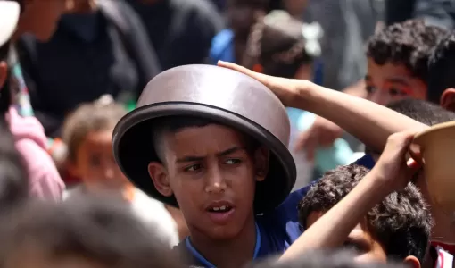 طفل فلسطيني يحمي رأسه من الشمس بوعاء في انتظار الحصول على طعام من تكية في مخيم النصيرات بقطاع غزة | المصدر: وكالة الأنباء الفرنسية