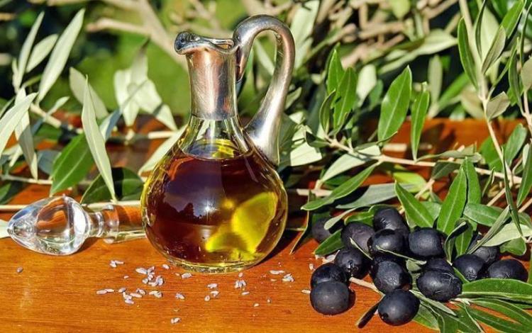 إبريق زجاجي مليء بزيت الزيتون وإلى جانبه ثمار زيتون سوداء وأغصان زيتون خضراء